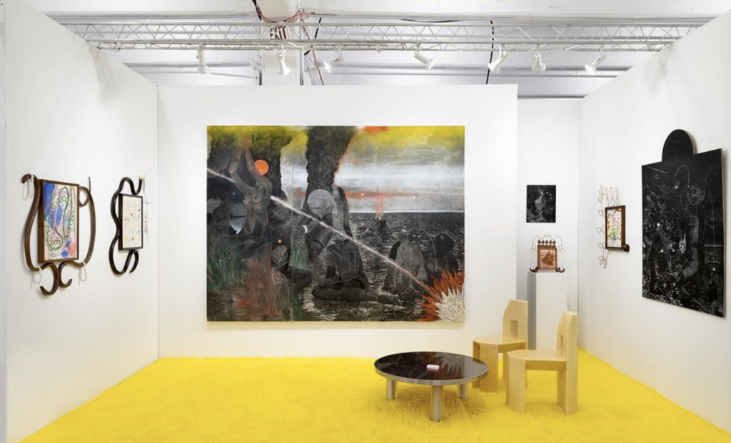 A color photo shows a white-walled booth with many paintings and a table and chair sitting on a yellow floor